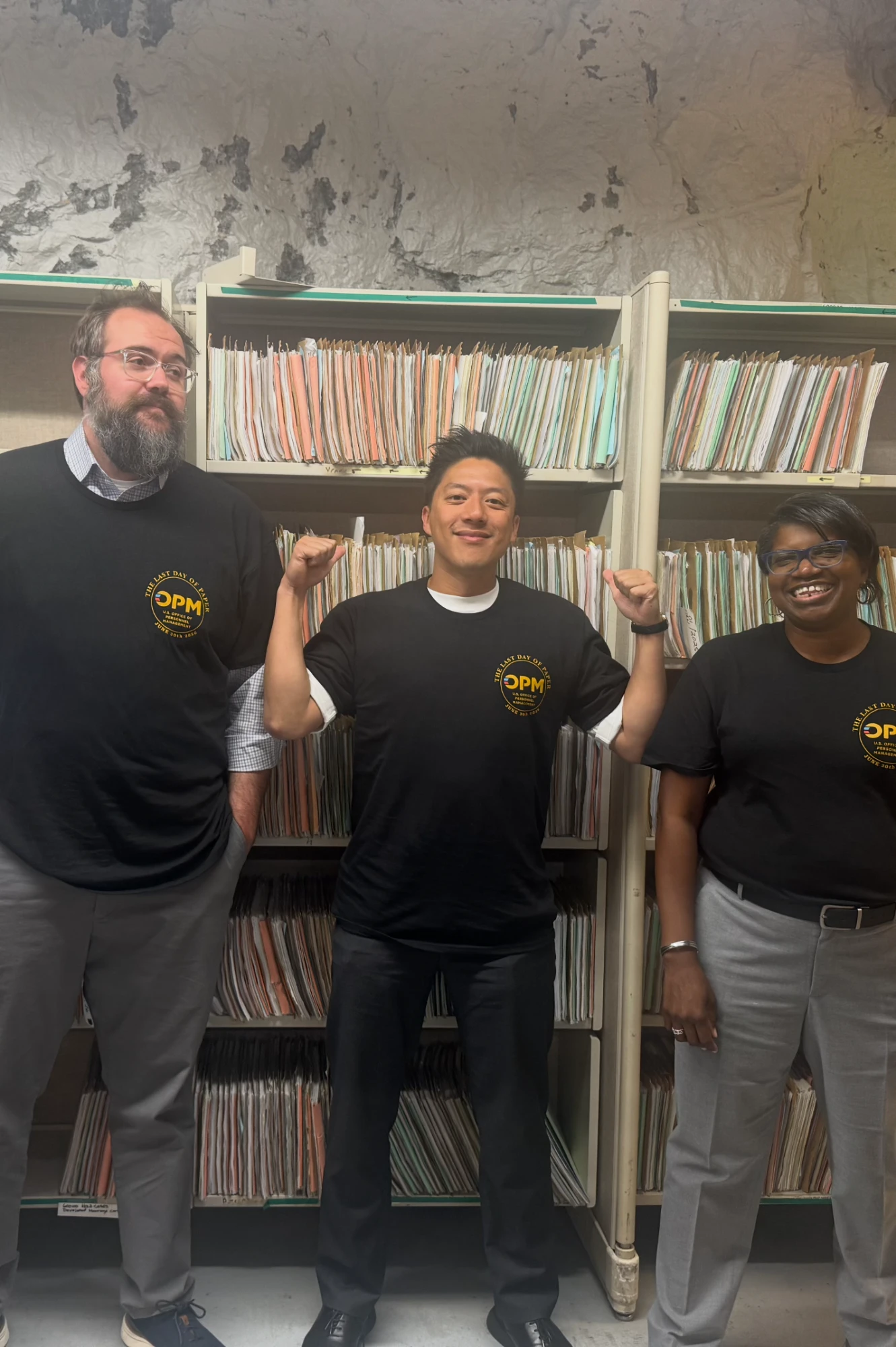 Three team members in “Last Day of Paper” shirts celebrating in front of shelves of paper retirement files